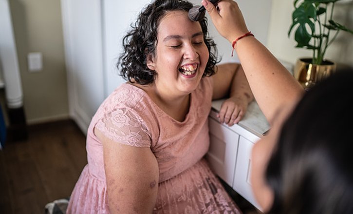 A support worker applying makeup with a brush to a young lady's face, both sharing a joyful and pleasant moment.