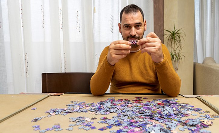 Man at table, concentrating on fitting puzzle piece into large scattered puzzle.